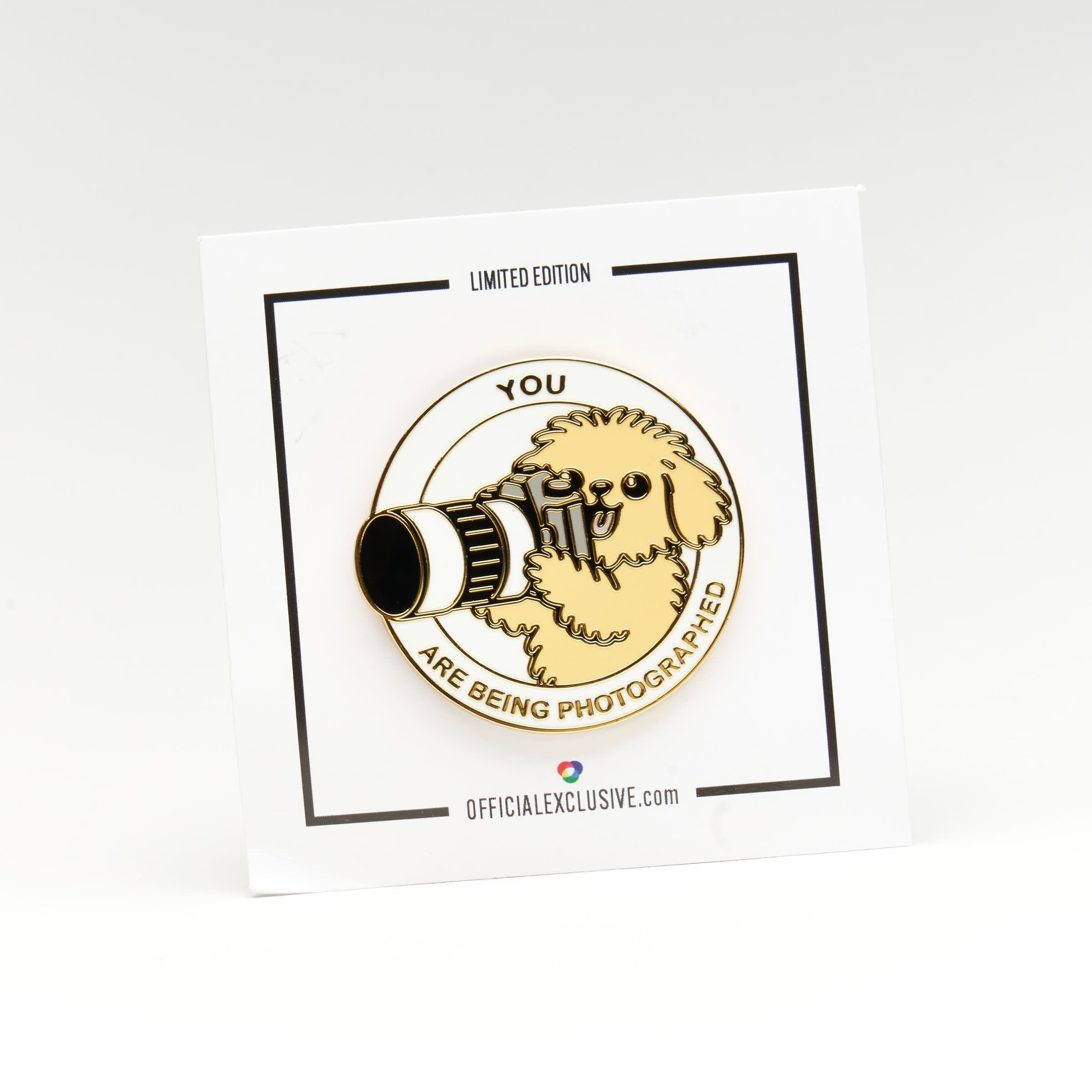 A circular enamel pin featuring a small dog holding a camera, with the text 'YOU ARE BEING PHOTOGRAPHED' around it, mounted on a white display card. The card has black border lines and text reading 'LIMITED EDITION' and 'OFFICIALEXCLUSIVE.