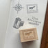 A wooden rubber stamp with an engraved postage stamp design reading 'AIR MAIL' and an airplane motif, placed on an open notebook. The notebook shows matching stamped designs, including a compass, an airplane mail stamp, a film canister, a pencil, and the text 'live Analogue'.