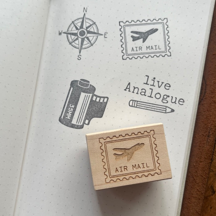 A wooden rubber stamp with an engraved postage stamp design reading 'AIR MAIL' and an airplane motif, placed on an open notebook. The notebook shows matching stamped designs, including a compass, an airplane mail stamp, a film canister, a pencil, and the text 'live Analogue'.