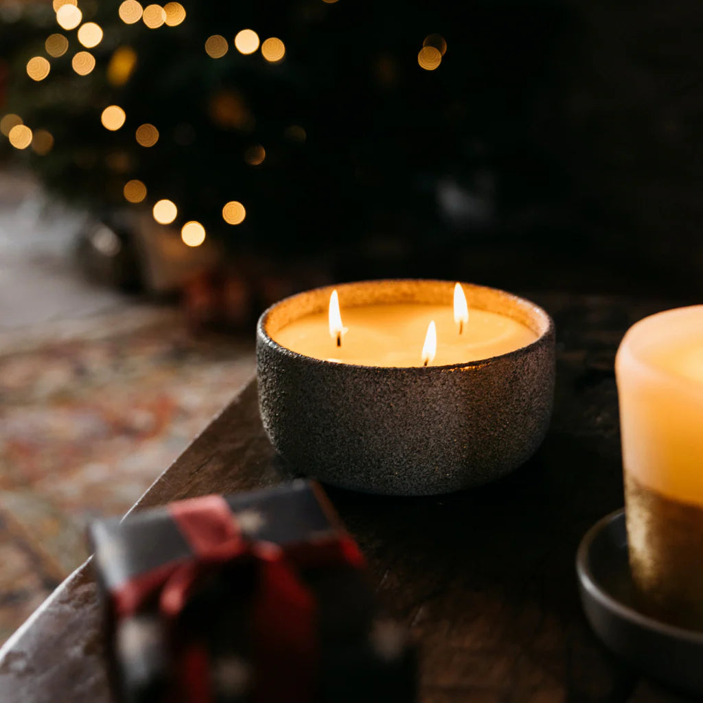Two lit candles in textured holders on a wooden surface with blurred lights in the background