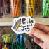Hand holding a 'Pride and Joy' sticker with colorful design in front of stationery items.