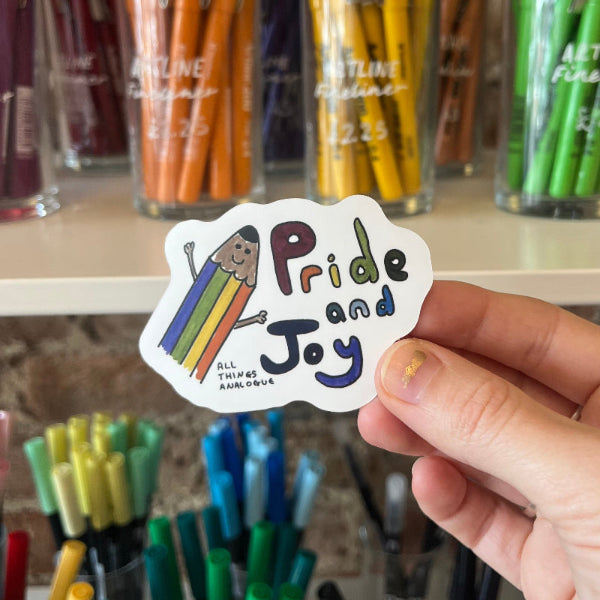 Hand holding a 'Pride and Joy' sticker with colorful design in front of stationery items.