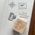 A wooden rubber stamp with an engraved film can design rests on an open notebook page showing stamped travel-themed designs, including a compass rose, an air mail stamp, and the words 'live Analogue'.