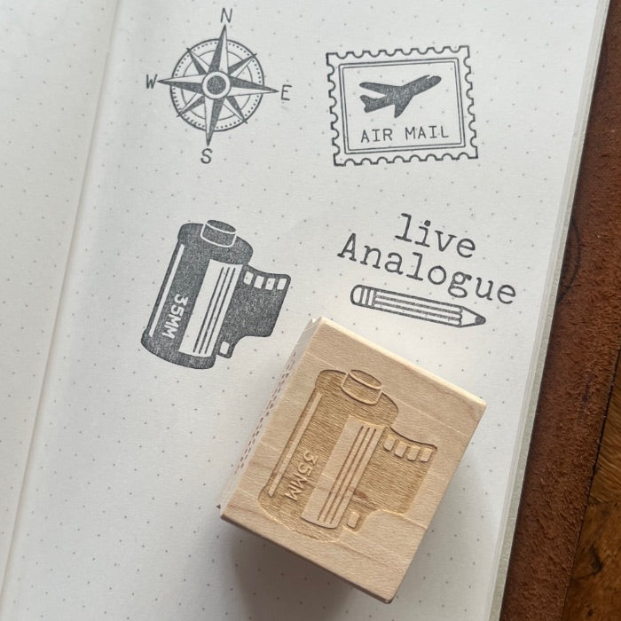 A wooden rubber stamp with an engraved film can design rests on an open notebook page showing stamped travel-themed designs, including a compass rose, an air mail stamp, and the words 'live Analogue'.