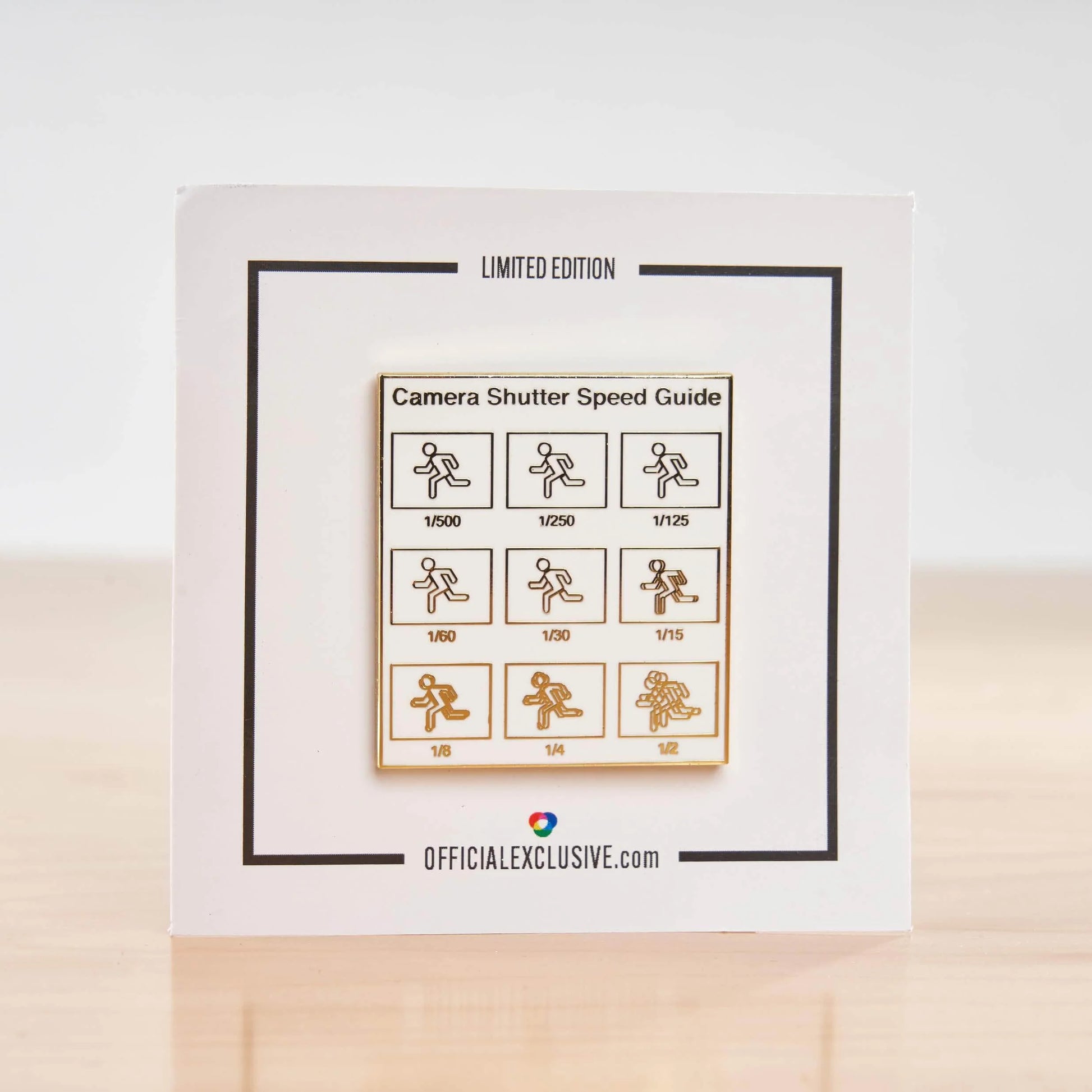 A square white card featuring a 'Camera Shutter Speed Guide' with black border lines and a central gold-bordered panel showing shutter speeds as icons. The card is displayed upright on a light wooden surface against a white background.