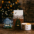 Two candles with decorative boxes on a wooden surface in front of a Christmas tree.