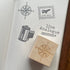 A wooden rubber stamp with a compass rose design rests on an open notebook page showing stamped black illustrations, including a compass, an air mail stamp, a film canister, and the words 'live Analogue'.