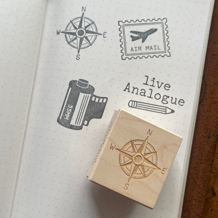 A wooden rubber stamp with a compass rose design rests on an open notebook page showing stamped black illustrations, including a compass, an air mail stamp, a film canister, and the words 'live Analogue'.