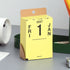 A yellow desk calendar page standing upright on a gray tabletop, with brown cardboard hanger tabs and black printed text showing 2027, Friday, 1, and Jan. Surrounding objects include a white cup, a green container, and office supplies.
