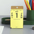 A yellow Penco 2027 desk calendar showing the 1st of January, standing on a gray desk with office supplies in the background.