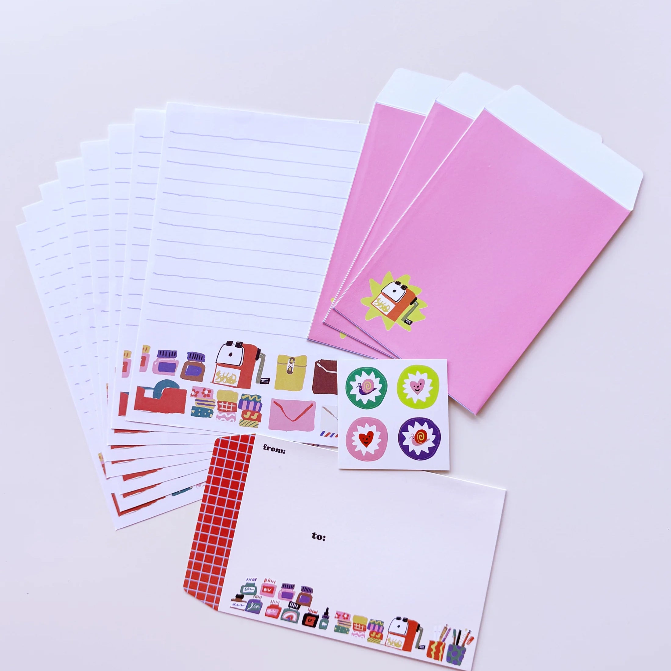 A stationery set arranged on a light background, including lined paper envelopes, pink card sleeves, sticker sheets with colorful icons, and a white letterhead card with a red checkered border.
