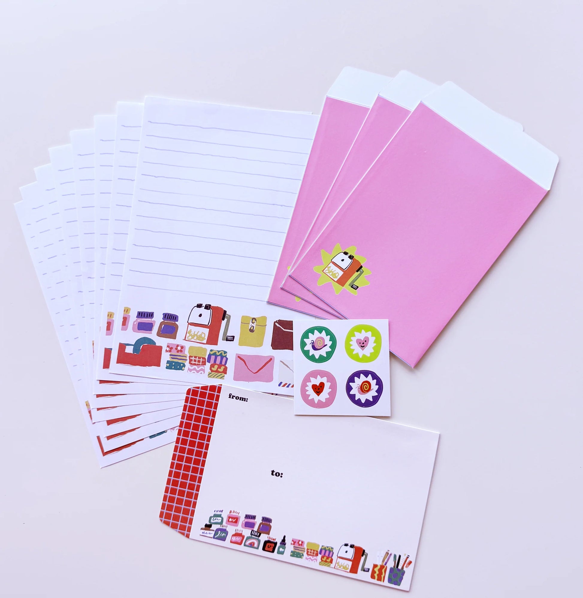 A stationery set arranged on a light background, including lined paper envelopes, pink card sleeves, sticker sheets with colorful icons, and a white letterhead card with a red checkered border.