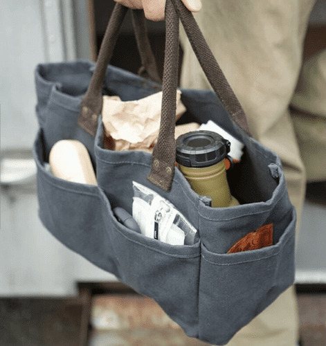 A dark gray canvas utility tote bag with brown handles, shown being carried by a person. The bag is filled with various items, including a beige bottle, a green travel mug, and small accessories, against a softly blurred outdoor background.