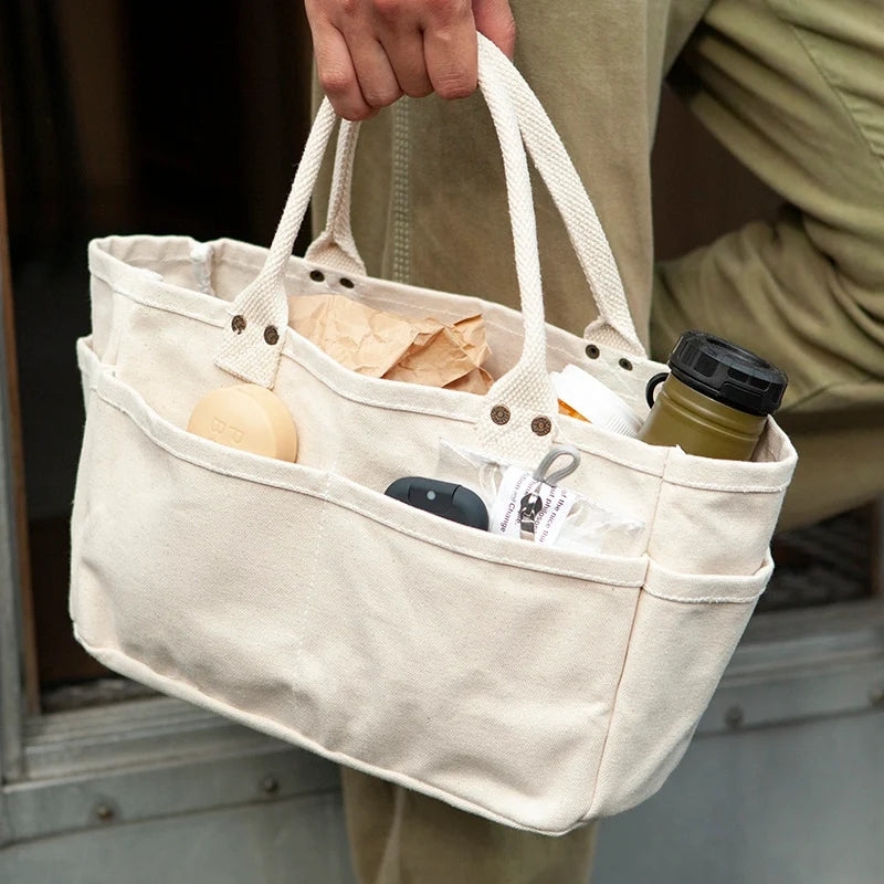 A person holds a large off-white canvas tote bag with multiple compartments and top handles. The bag contains everyday items like a brown paper bag, a round beige item, a dark case, and a green bottle.