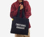 Person holding a black tote bag with 'EMOTIONAL BAGGAGE' text on a white background