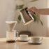 Person pouring coffee from a stainless steel kettle into a glass carafe on a wooden table.