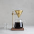 Coffee brewing setup with glass carafe, brass dripper, and wooden stand on a gray background