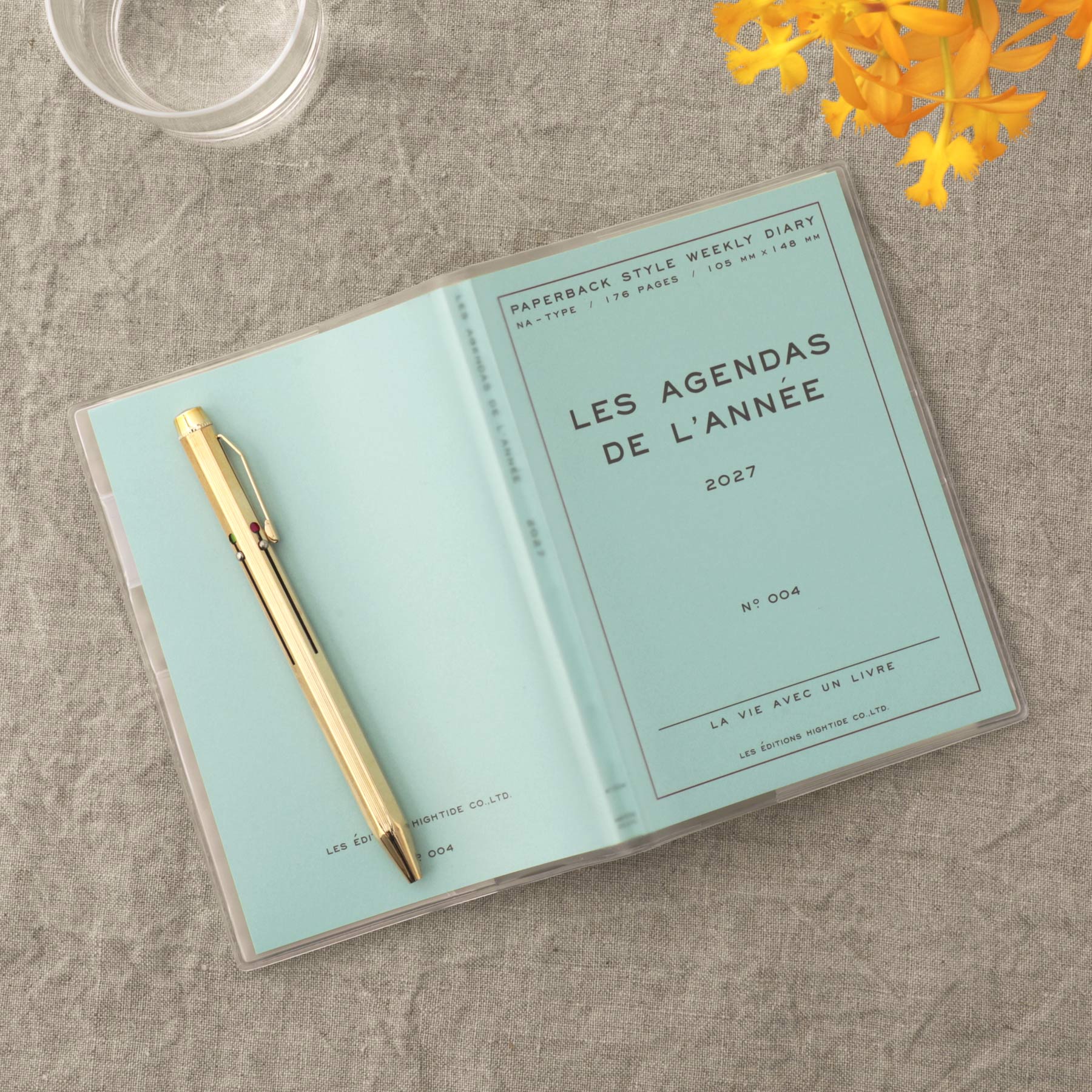 A clear plastic sleeve holding a light blue yearly planner and a gold pen, photographed on a textured gray fabric surface with a glass and yellow flowers as props.