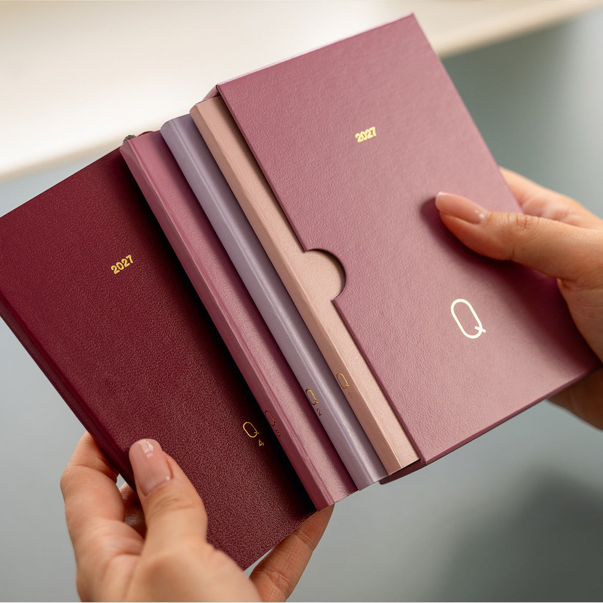 Four slim 2027 pocket calendars in burgundy, mauve, dusty pink, and lavender, held in two hands to show the thickness and texture of the covers.