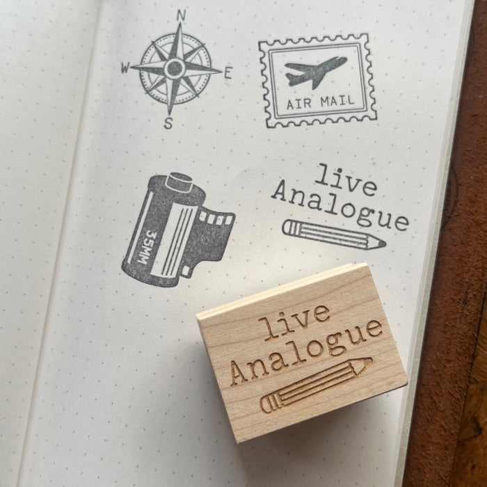 A wooden rubber stamp with the engraved text 'live Analogue' and a pencil illustration, resting on an open dotted notebook that shows matching stamped designs including a compass, postage stamp, film canister, and the phrase 'live Analogue'.