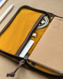 A pocket journal with a waxed canvas cover, displayed alongside a pen and cables, revealing the interior dot grid refill and organization pockets.