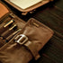 A partially unrolled tool roll with a brown waxed canvas exterior, aluminum hook fastener, and multiple pockets containing pens and other tools.