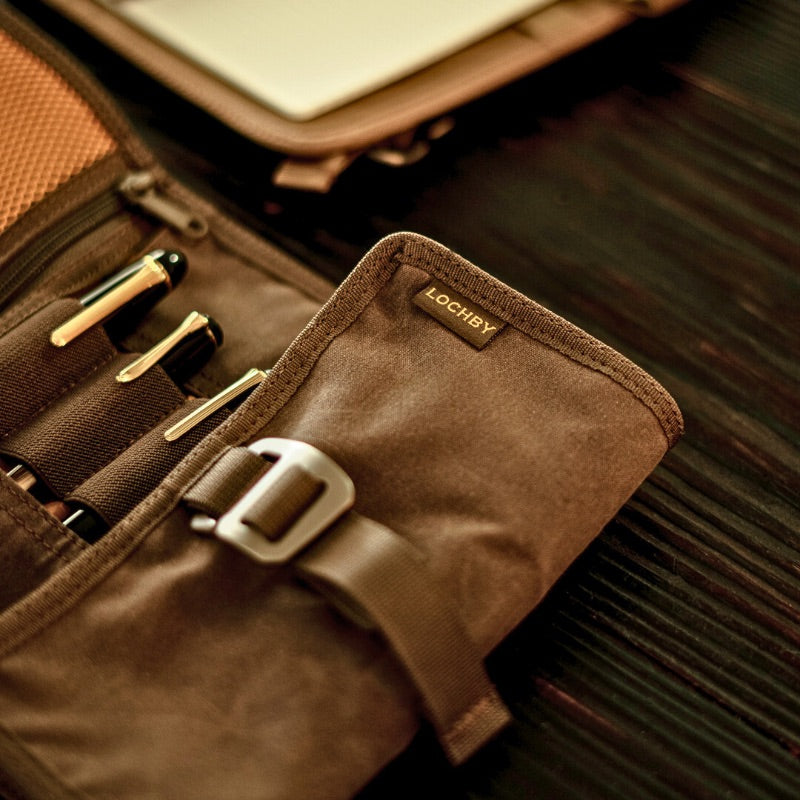 A partially unrolled tool roll with a brown waxed canvas exterior, aluminum hook fastener, and multiple pockets containing pens and other tools.