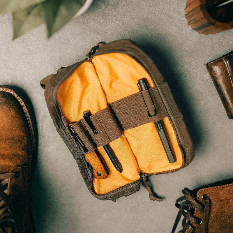 A brown and yellow venture pouch with elastic loops and pockets, alongside a pair of shoes and a bag, suggesting it can be carried in a bag or used as a crossbody bag.
