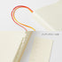 An off-white leather-bound notebook shown with its matching tabbed index page and numbered index stickers, featuring red and orange ribbon bookmarks on a clean white background.