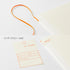 A cream-colored library card organizer box with an orange loop handle and matching orange printed details, shown alongside a sample library card insert with Japanese text on a white background.