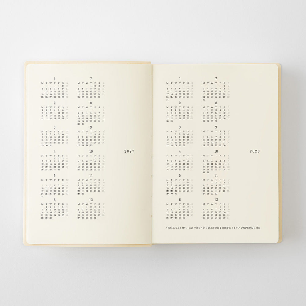 An open cream-colored planner or notebook showing printed calendar pages with dates and Japanese text, photographed against a light gray background.