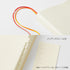 An off-white notebook or journal shown in a split view: the top half shows a close-up of the textured cover with two ribbon bookmarks in red and orange, and the bottom half shows the open interior with page tabs and a page insert labeled with page numbers. Japanese text appears on the right.