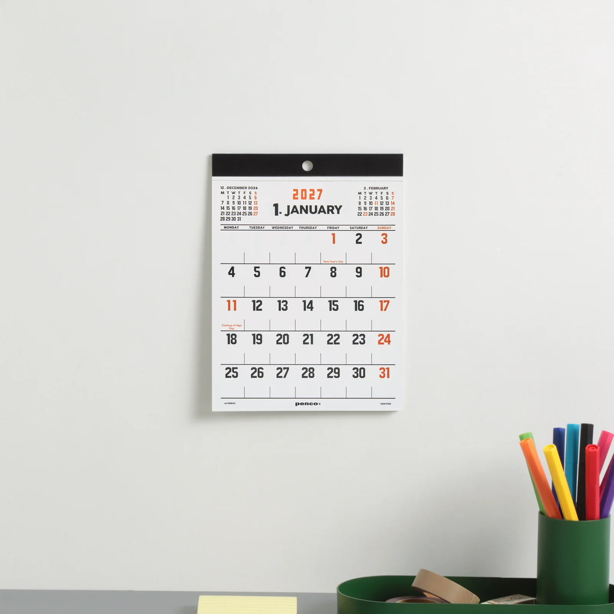 A 2027 January wall calendar hanging on a light gray wall above a desk with a green cup of colored pencils.