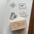 A wooden rubber stamp with an engraved postage stamp design rests on the pages of a dotted notebook. The notebook shows several stamp impressions, including a compass rose, an air mail stamp, a film can stamp, a pencil, and the words 'live Analogue'.