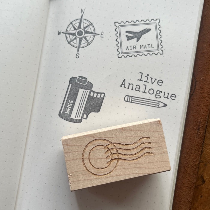 A wooden rubber stamp with an engraved postage stamp design rests on the pages of a dotted notebook. The notebook shows several stamp impressions, including a compass rose, an air mail stamp, a film can stamp, a pencil, and the words 'live Analogue'.