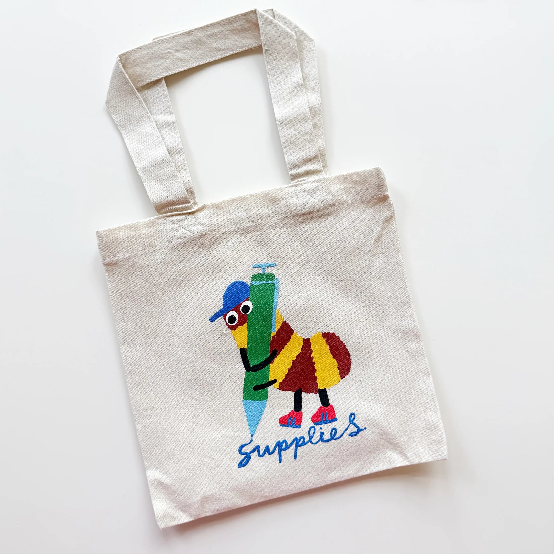 A natural canvas tote bag with a colorful cartoon bee holding a green pencil and the word 'Supplies' printed in blue script, shown on a plain white background.