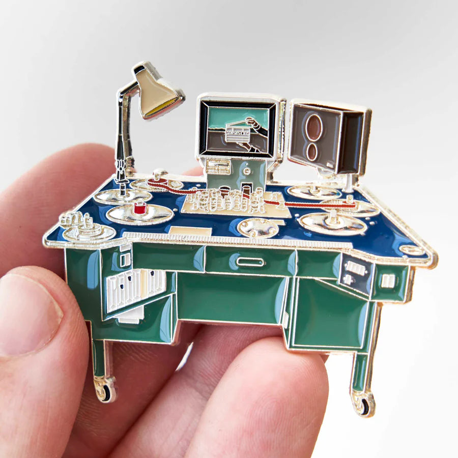 A miniature desk-themed enamel pin held in a person's hand against a light gray background. The pin features a blue desk with green drawers, gold-tone metal outlines, a silver desk lamp, a computer monitor, and a speaker.