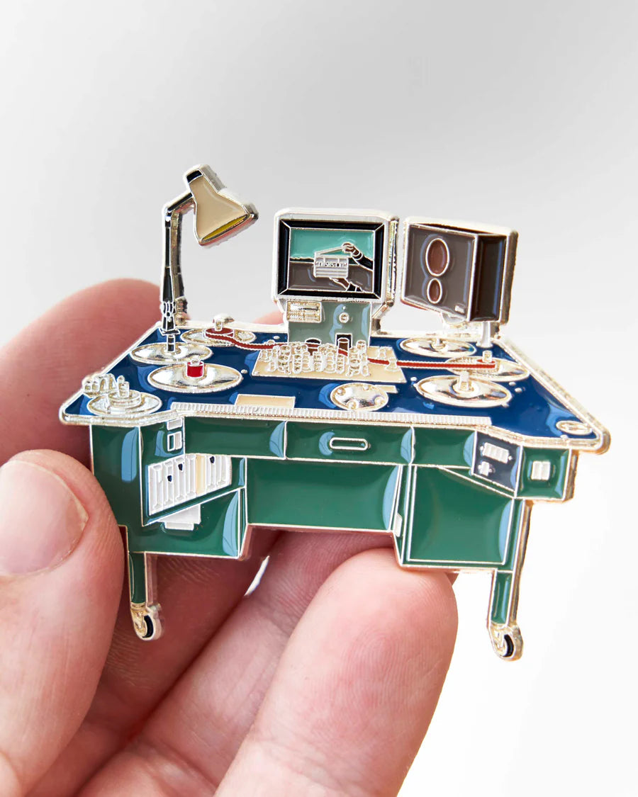 A miniature desk-themed enamel pin held in a person's hand against a light gray background. The pin features a blue desk with green drawers, gold-tone metal outlines, a silver desk lamp, a computer monitor, and a speaker.