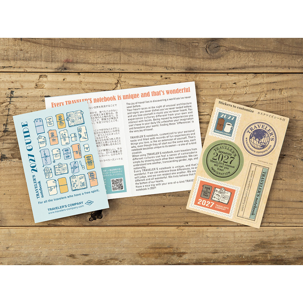 A flat-lay of a travel notebook set on a rustic wooden background, featuring a light blue 'Travel Guide' booklet, an open notebook with red and black text, and a cream-colored stamp booklet with colorful stamps.