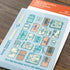 A sheet of decorative stickers featuring illustrated travel backpacks and passport-style holders in pastel blue, peach, cream, and teal, placed on a wooden surface. The sheet includes the text 'TRAVELER'S notebook' and 'PASSPORT SIZE CLEAR FOLDER' on an orange header, with the brand mark 'TRAVELER'S COMPANY' visible.