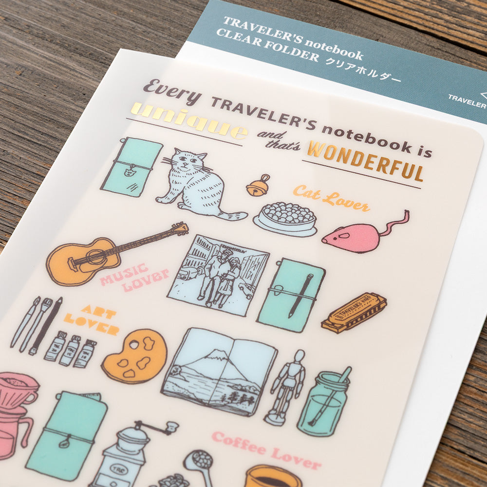 A clear folder printed with cute travel-themed illustrations and the phrase 'Every TRAVELER'S notebook is Wonderful' lies on a wooden surface. The folder has pastel drawings of a cat, guitar, mason jar, coffee maker, camera, and other travel motifs, with a teal header card visible behind it.