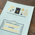 A light blue envelope-style stationery piece laid on a dark wood surface, printed with gold and black travel-themed illustrations and the phrase, 'Every traveler's notebook is unique and that's wonderful.'.