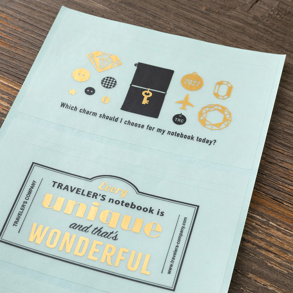 A light blue envelope-style stationery piece laid on a dark wood surface, printed with gold and black travel-themed illustrations and the phrase, 'Every traveler's notebook is unique and that's wonderful.'.