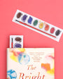 Colorful book with watercolor design, small paint palette, and larger paint palette on a pink background