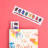 Colorful book with watercolor design, small paint palette, and larger paint palette on a pink background