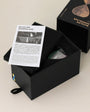 Black box containing a bicolor sandglass with an open lid and product information sheet.