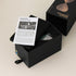 Black box containing a bicolor sandglass with an open lid and product information sheet.
