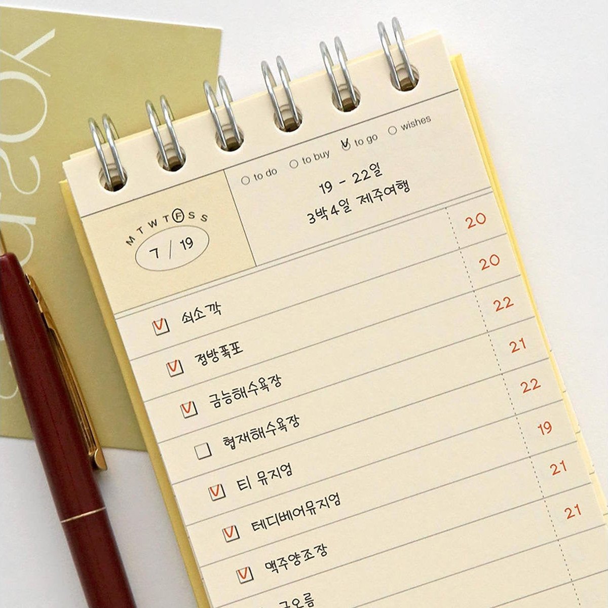 A spiral-bound planner or notebook page with a checklist layout in Korean, photographed on a light background with part of a gold card and a dark red pen visible.