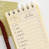 A spiral-bound planner or notebook page with a checklist layout in Korean, photographed on a light background with part of a gold card and a dark red pen visible.
