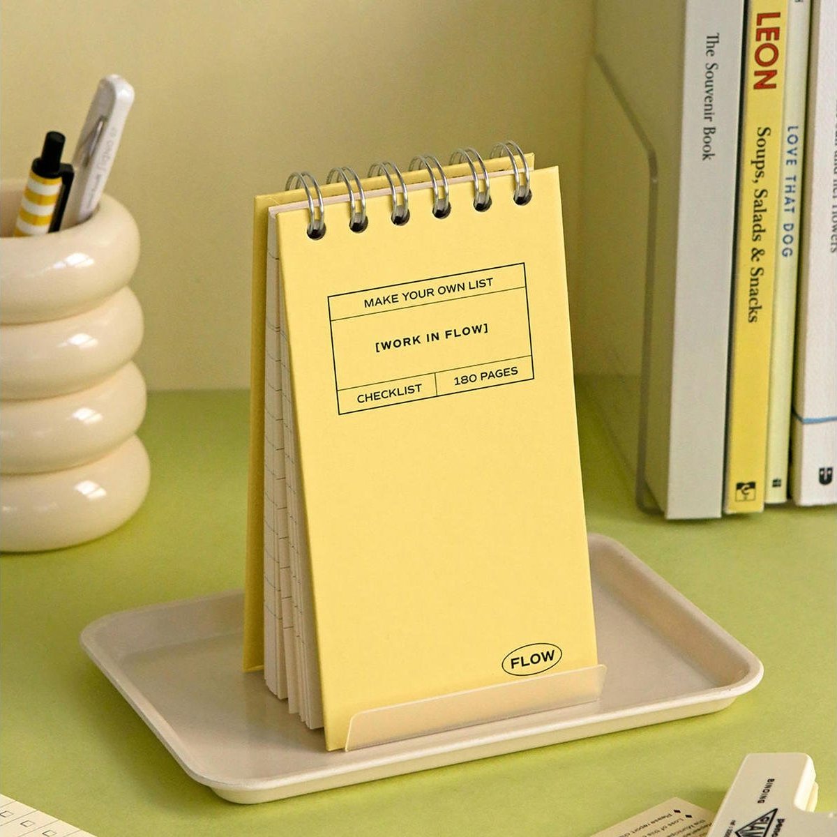 A yellow spiral-bound notepad displayed upright in a white holder on a tray, with a pen cup and books in the background.
